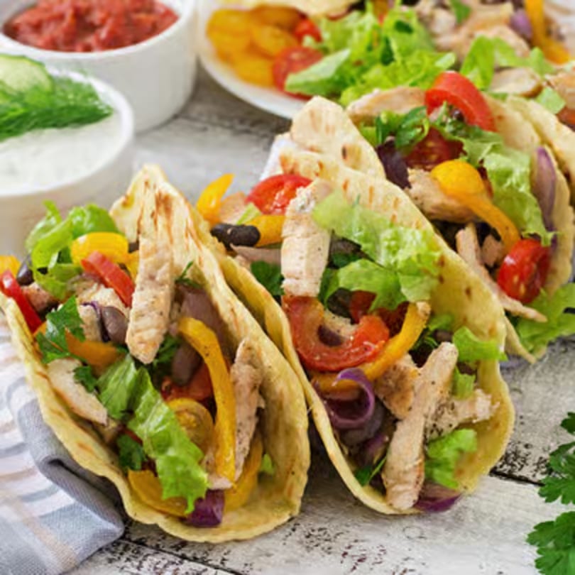 Three chicken tacos with colorful vegetables, salsa, and sour cream on a rustic wooden table.