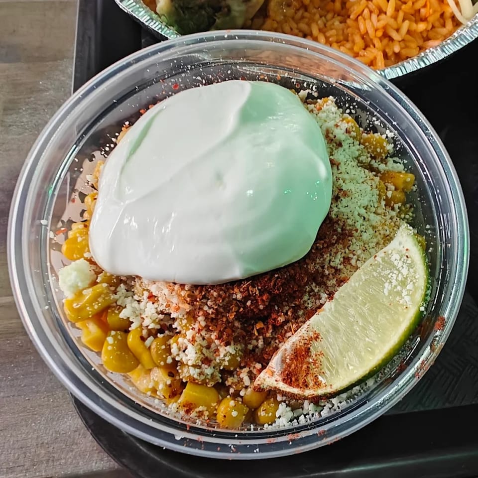 Mexican street corn (Esquites) in a clear bowl, topped with crema, chili powder, cheese, and a lime wedge.