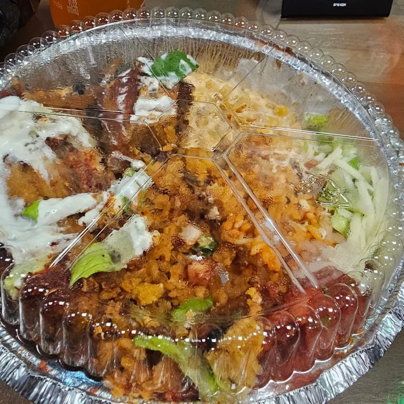 A foil container of Mexican food with rice, meat, and toppings, covered with a clear plastic lid.